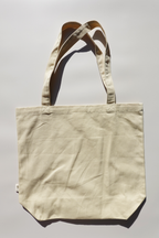 Plain beige canvas tote bag displayed flat on a light background under direct sunlight, showing the bag's texture, stitching, and structure.