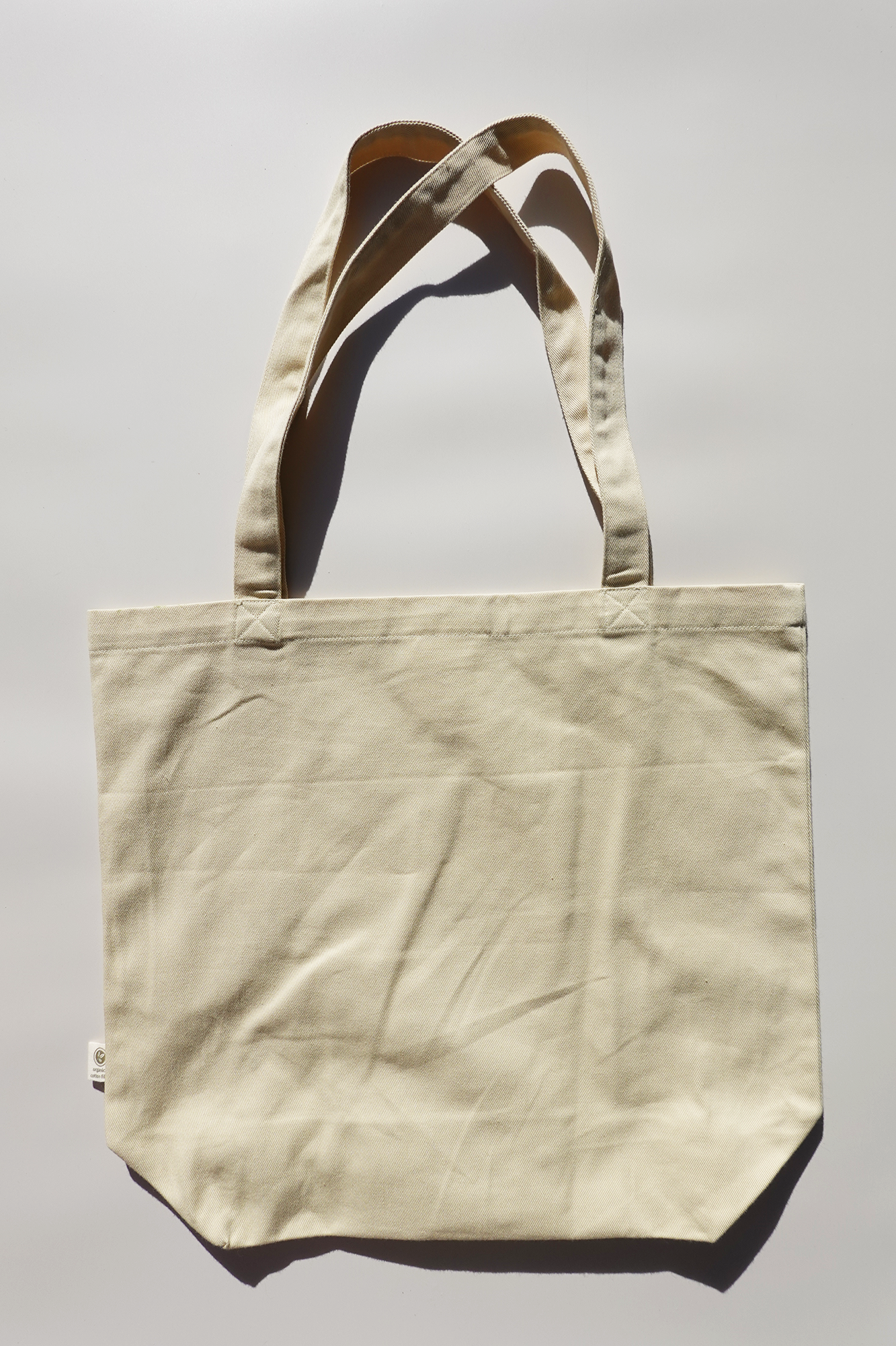 Plain beige canvas tote bag displayed flat on a light background under direct sunlight, showing the bag's texture, stitching, and structure.
