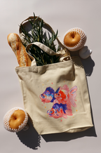 Beige canvas tote bag featuring a colorful illustration of three goldfish with blue and orange accents, surrounded by small stars. The bag is filled with greenery and a baguette, and is placed next to two foam-netted Asian pears and a small white bunny figurine on a light background.