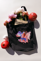Black canvas tote bag featuring an illustration of three colorful goldfish surrounded by small sparkles and stars. The bag is filled with peonies and a book titled 'Starting Point,' with pomegranates and a small white figurine placed around it on a light background.