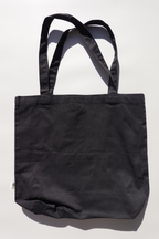 Plain black eco cotton tote bag laid flat on a light background under natural sunlight, shown empty to highlight shape, structure, and size — reusable and minimalist bag ideal for everyday errands, books, or custom designs