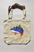 Beige canvas tote bag featuring an illustration of a blueberry tart topped with cute cartoon bunny characters, centered on a plain background under direct light.