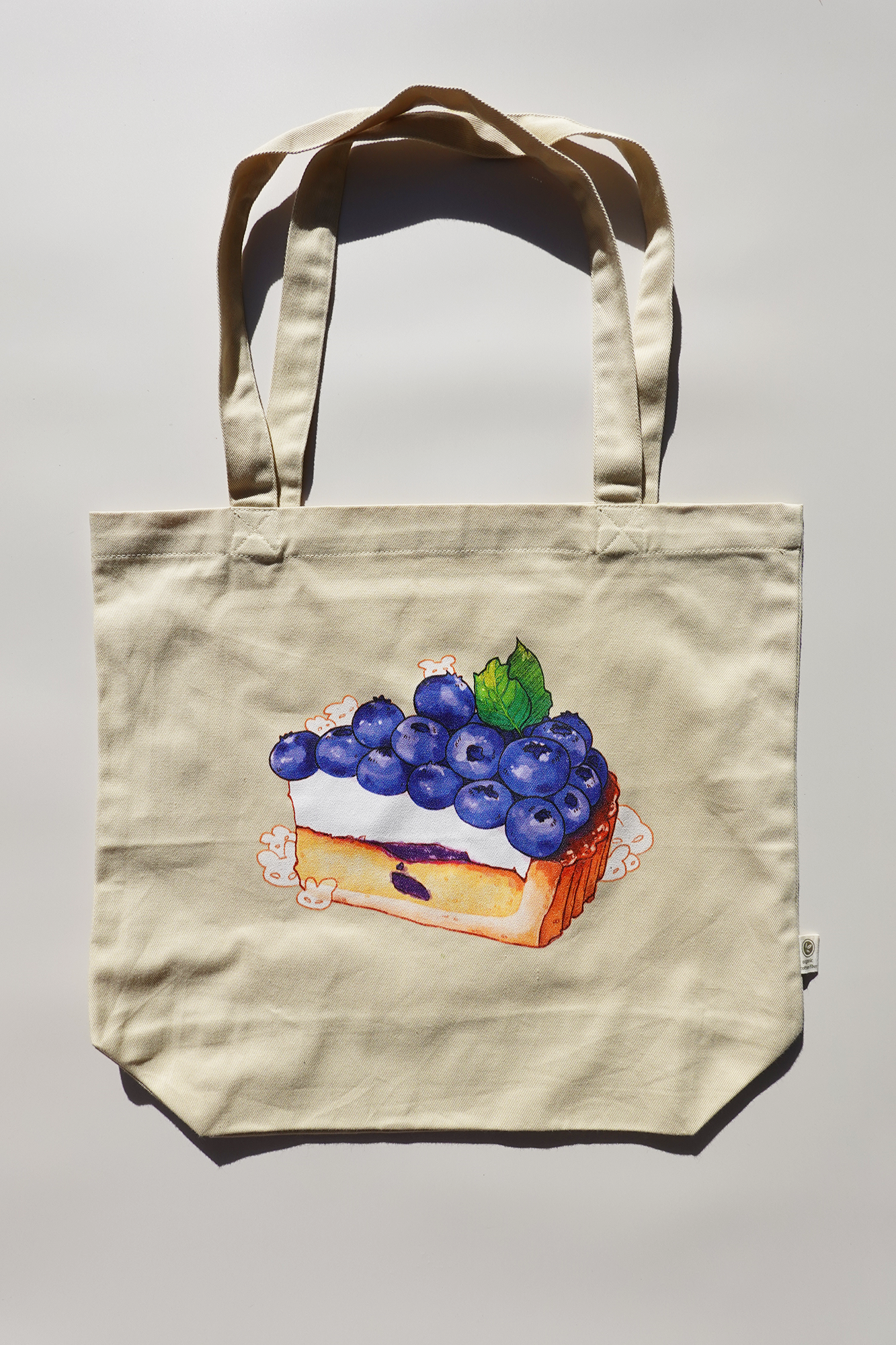 Beige canvas tote bag featuring an illustration of a blueberry tart topped with cute cartoon bunny characters, centered on a plain background under direct light.