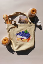 Beige canvas tote bag with a vibrant illustration of a blueberry tart topped with small white cartoon bunnies, styled with a baguette, postcard prints, and Asian pears in mesh wraps, under bright natural light.