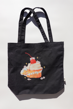Black canvas tote bag featuring an illustration of a cupcake topped with whipped cream and a cherry, surrounded by small white bunnies and sparkles. The bag is photographed flat against a light background with strong natural shadows.
