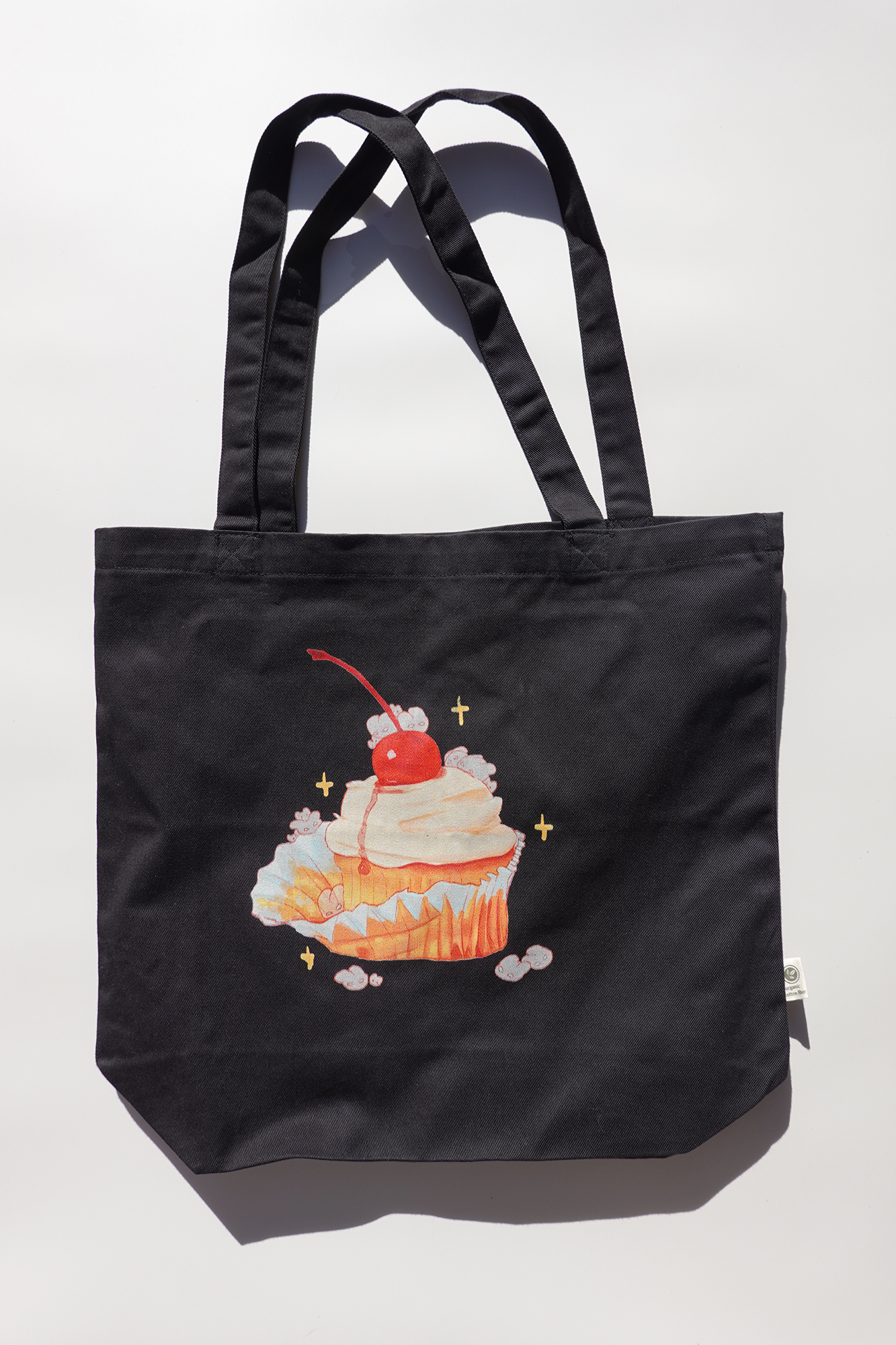 Black canvas tote bag featuring an illustration of a cupcake topped with whipped cream and a cherry, surrounded by small white bunnies and sparkles. The bag is photographed flat against a light background with strong natural shadows.