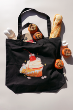 Black canvas tote bag featuring an illustration of a whipped cream cupcake with a cherry on top, surrounded by tiny white bunnies and sparkles. The tote is filled with a baguette, persimmons, a Hayao Miyazaki book, and a white bunny-shaped bottle, styled against a bright backdrop with shadows.