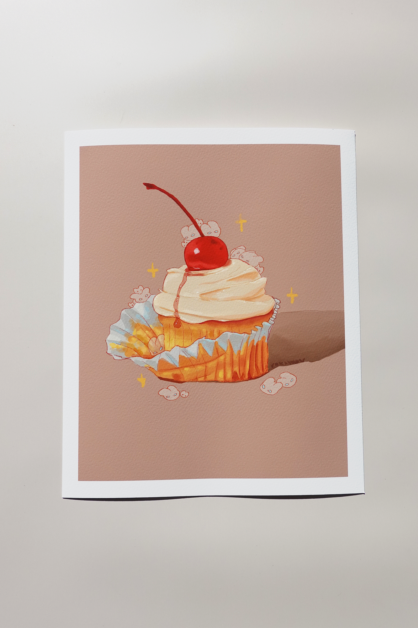 Fine art print of a cherry-topped cupcake illustration with whipped cream, surrounded by hidden bunny characters and sparkles on a warm beige background — printed on textured paper and displayed under soft studio lighting