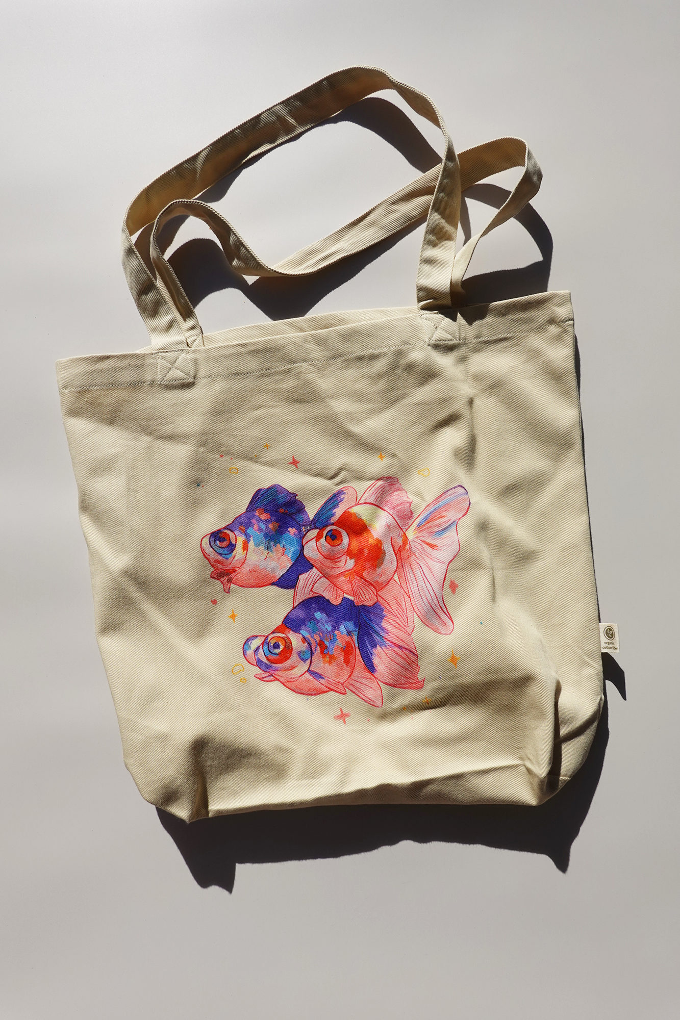 Beige canvas tote bag featuring a colorful illustration of three goldfish with vibrant blue and orange hues, surrounded by small stars. The bag is laid flat on a light surface under strong natural light, casting clear shadows.