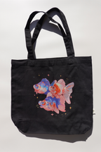 Black canvas tote bag featuring a colorful illustration of three goldfish with blue and orange accents, surrounded by small stars and sparkles on a plain light background.