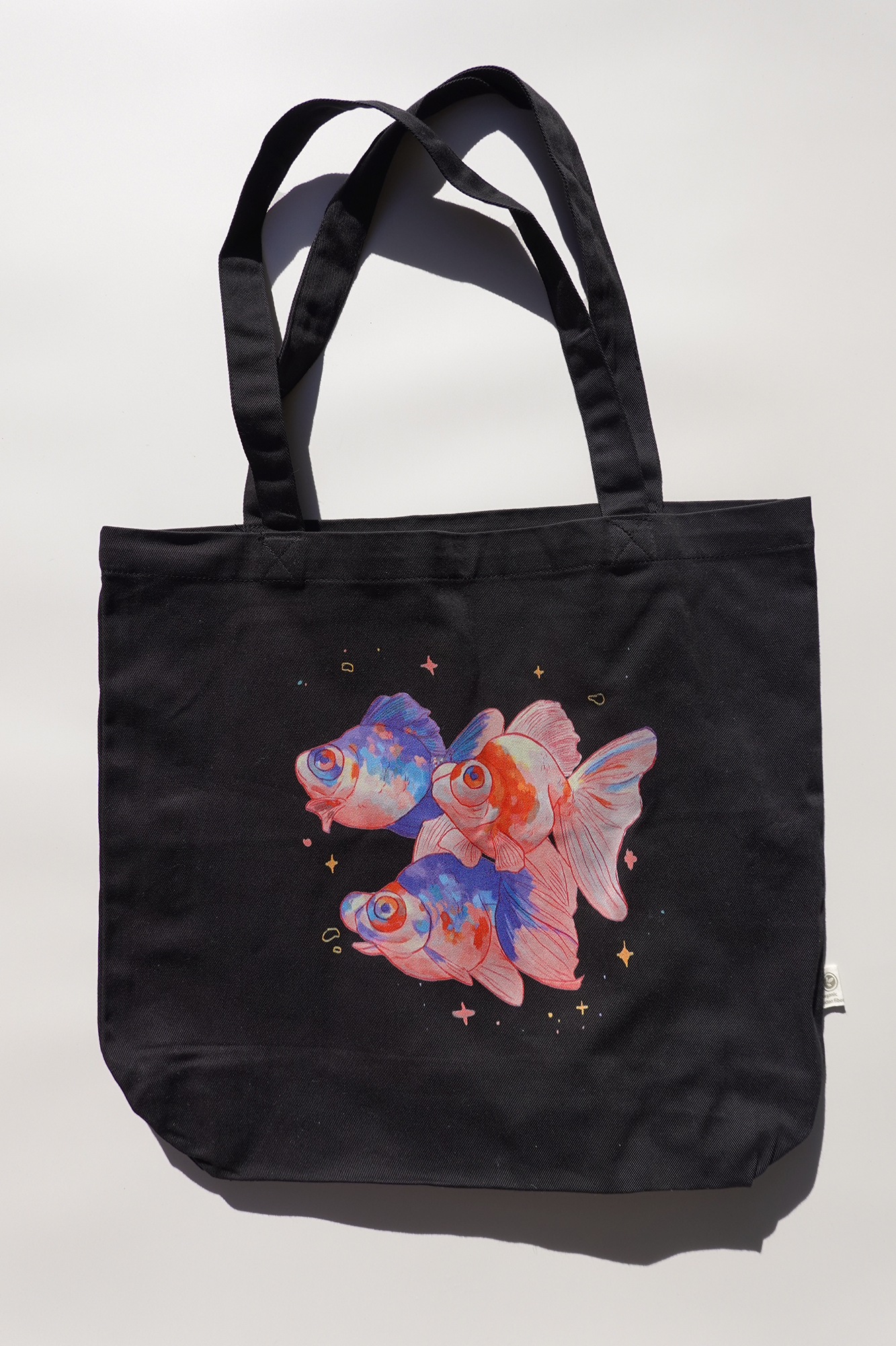 Black canvas tote bag featuring a colorful illustration of three goldfish with blue and orange accents, surrounded by small stars and sparkles on a plain light background.