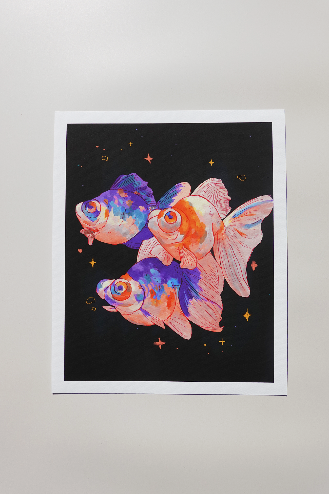 Art print of three colorful goldfish painted in vibrant orange, pink, and blue hues, swimming against a black starry background with glowing sparkles — printed on textured fine art paper and photographed in soft natural light