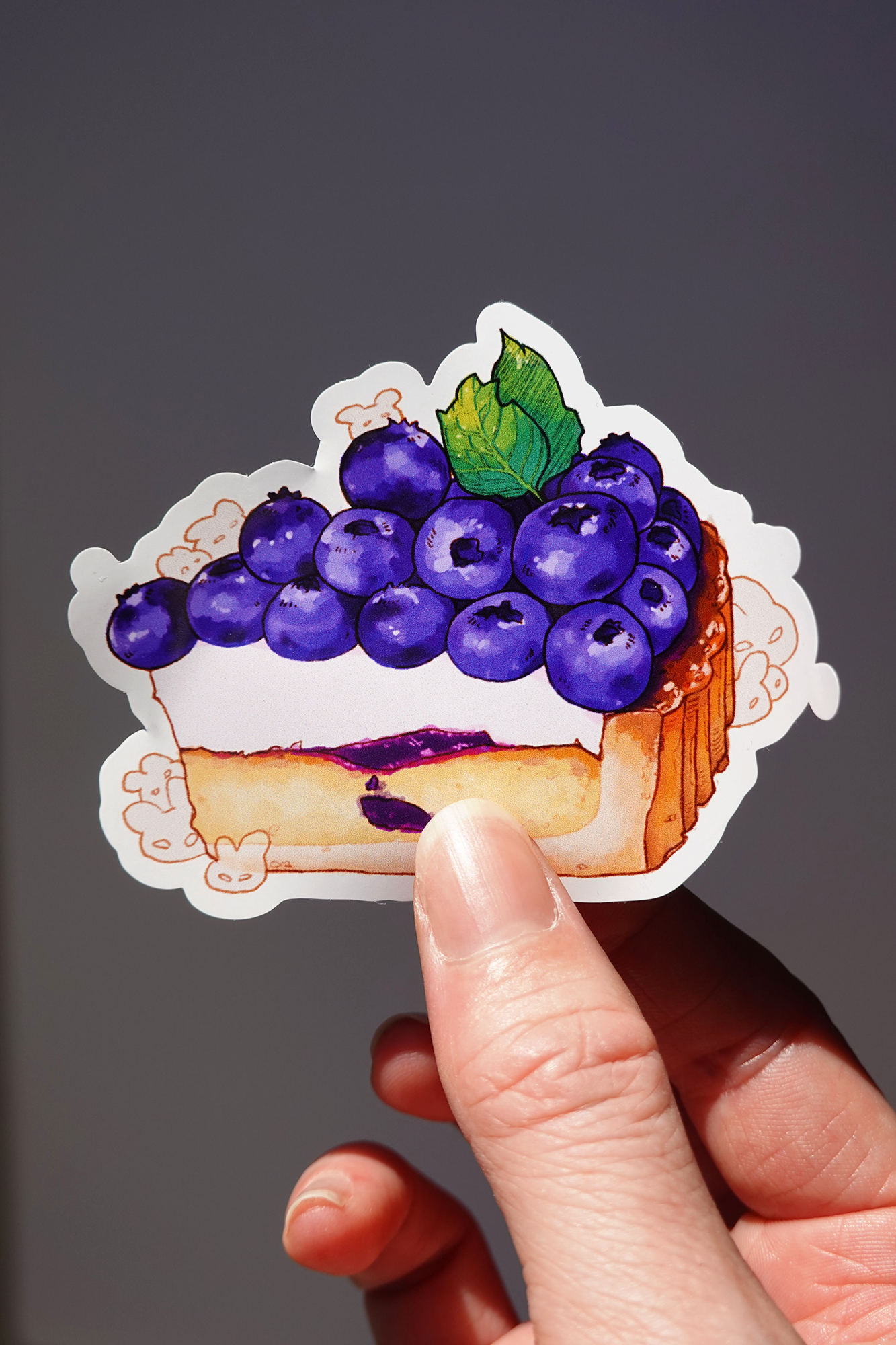 Blueberry cheesecake vinyl sticker featuring a slice topped with plump, glossy blueberries and a mint leaf, with hidden bunny characters around the base — held in hand against a neutral background, cute dessert-themed die-cut sticker for decorating journals, laptops, or water bottles
