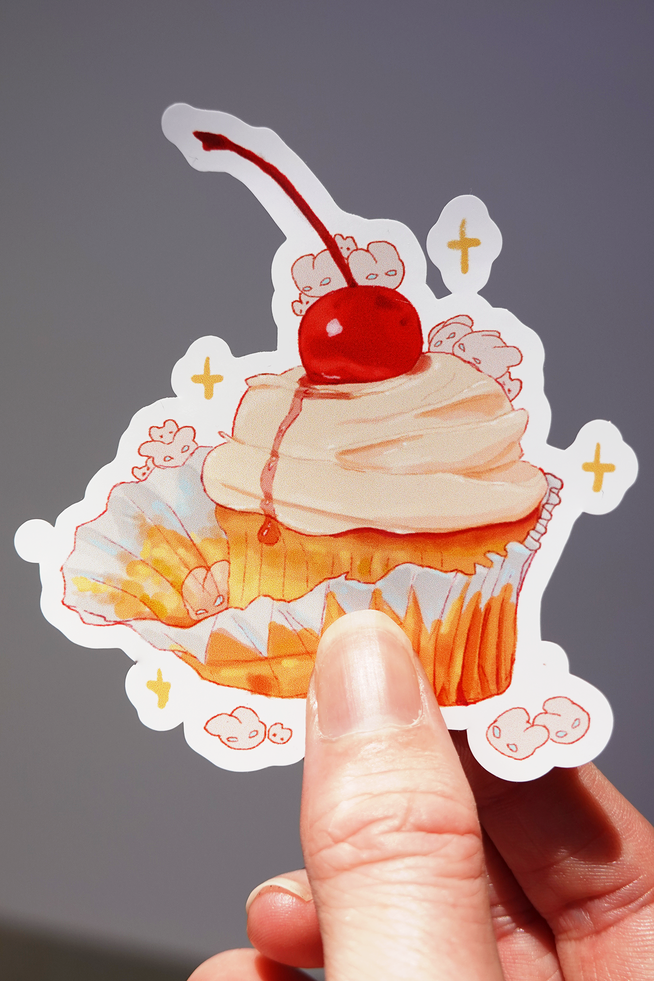 Cherry cupcake vinyl sticker featuring a golden vanilla cupcake with swirled frosting and a glossy red cherry on top, surrounded by hidden bunny characters and sparkles — held in hand against a neutral background, cute dessert-themed die-cut sticker for decorating water bottles, laptops, or journals