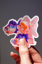 Colorful goldfish vinyl sticker featuring three vibrant orange, pink, and purple fish with expressive eyes and flowing fins — held in hand against a neutral background, glossy die-cut aquatic-themed art sticker for laptops, water bottles, or journals
