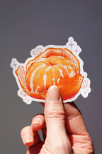 Peeled mandarin orange vinyl sticker with juicy orange segments and whimsical hidden bunny characters, illustrated in a vibrant hand-drawn style with purple sparkle accents — held in hand against a neutral background, cute fruit-themed die-cut sticker for journaling, laptops, or water bottles