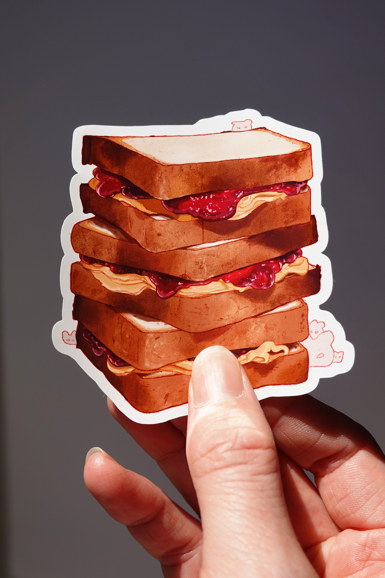 Peanut butter and jelly sandwich vinyl sticker featuring a whimsical stack of five thick-cut sandwiches with oozing fillings, illustrated in warm brown and berry tones with tiny hidden bunny characters — held in hand against a neutral background, glossy food-themed die-cut sticker for water bottles, laptops, or lunchbox decor