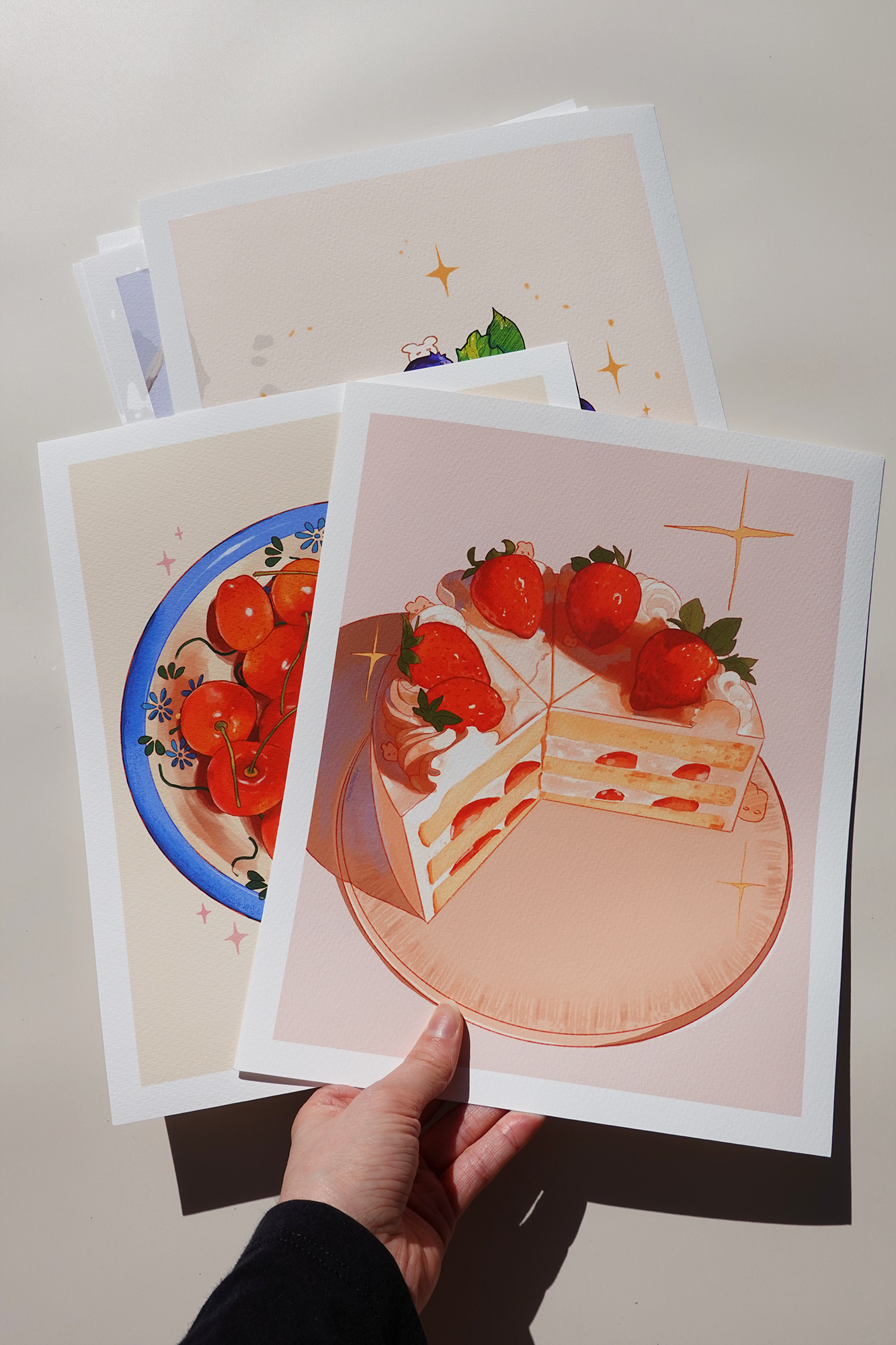 Hand holding a small stack of dessert-themed fine art prints featuring cherry bowl, strawberry shortcake, and blueberry cheesecake illustrations with hidden bunny characters — printed on textured paper and displayed under soft daylight on a neutral background