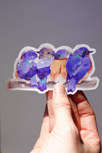 Bluebird vinyl sticker featuring four plump, colorful birds perched closely on a branch, illustrated in vibrant shades of blue, purple, and orange — held in hand against a neutral background, cute nature-inspired die-cut sticker for journaling, laptops, or water bottles