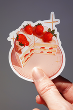 Strawberry shortcake vinyl sticker featuring a sliced layered cake topped with whole strawberries and whipped cream, with hidden bunny characters and sparkles around the plate — held in hand against a neutral background, cute dessert-themed die-cut sticker for journaling, laptops, or water bottles
