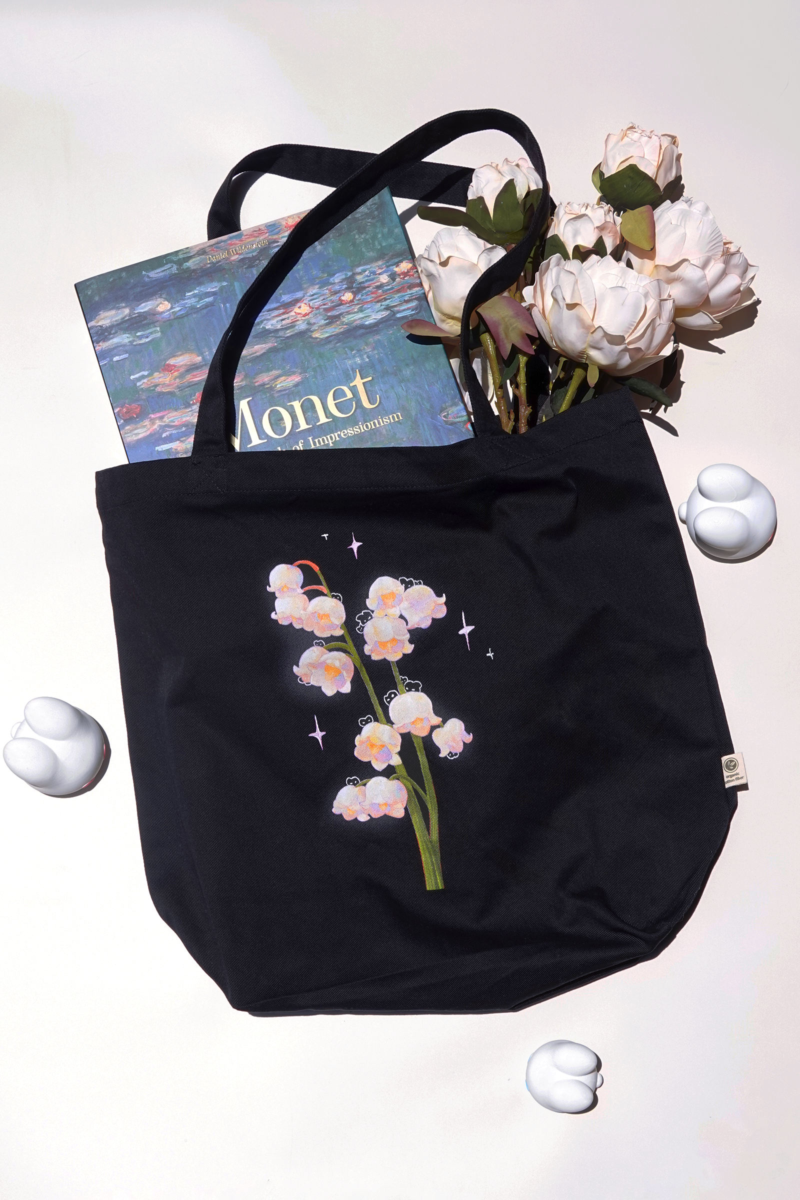 Black cotton tote bag featuring a lily of the valley floral design with hidden bunnies and pastel sparkles, styled with a Monet art book and soft pink roses — surrounded by white bunny-shaped figures on a light background, ideal for artists, book lovers, or floral stationery fans
