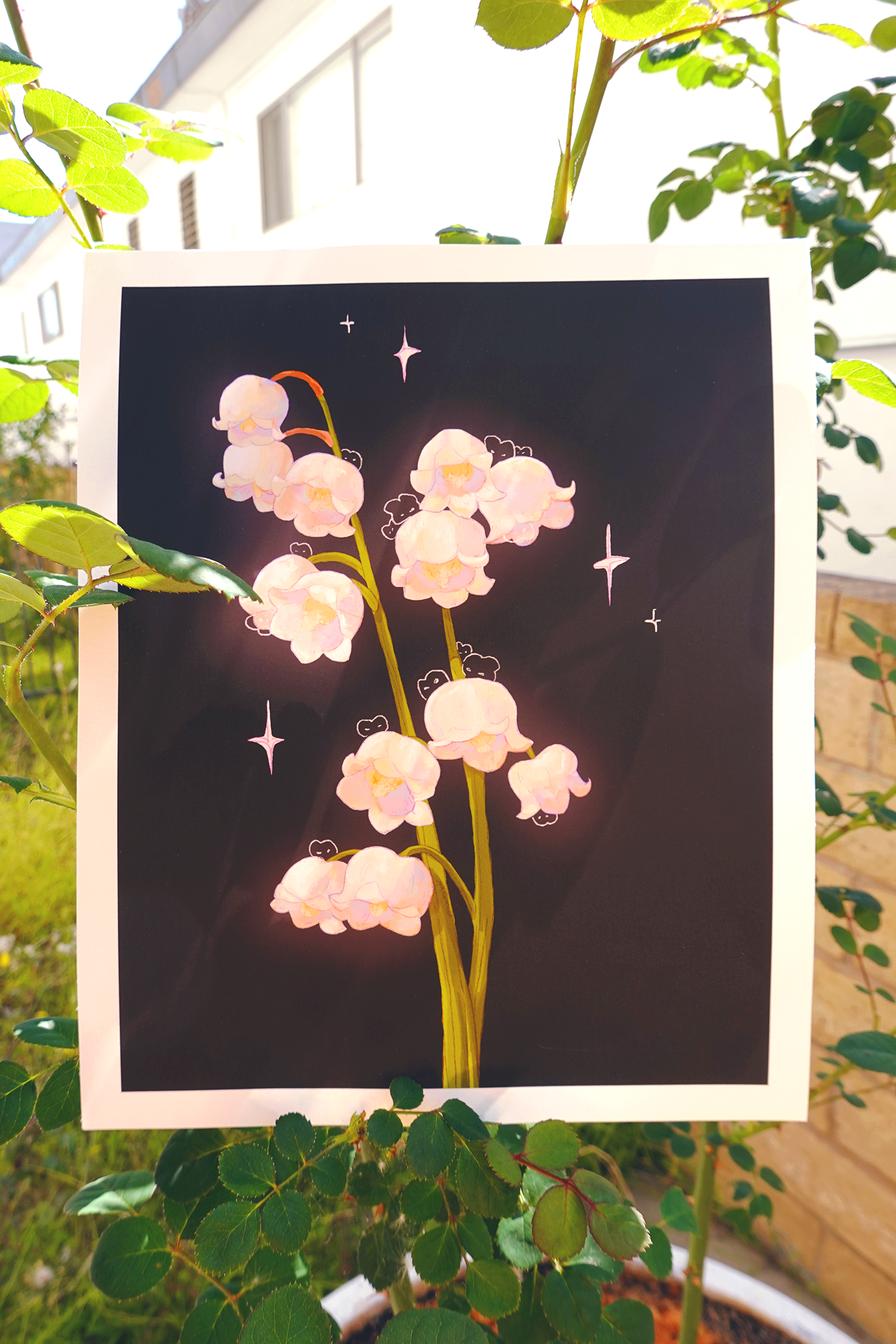 Lily of the valley fine art print with hidden bunnies and soft pastel sparkles on a black background, displayed outdoors among garden rose stems in natural sunlight — whimsical botanical illustration perfect for wall decor, cottagecore rooms, or nature lovers