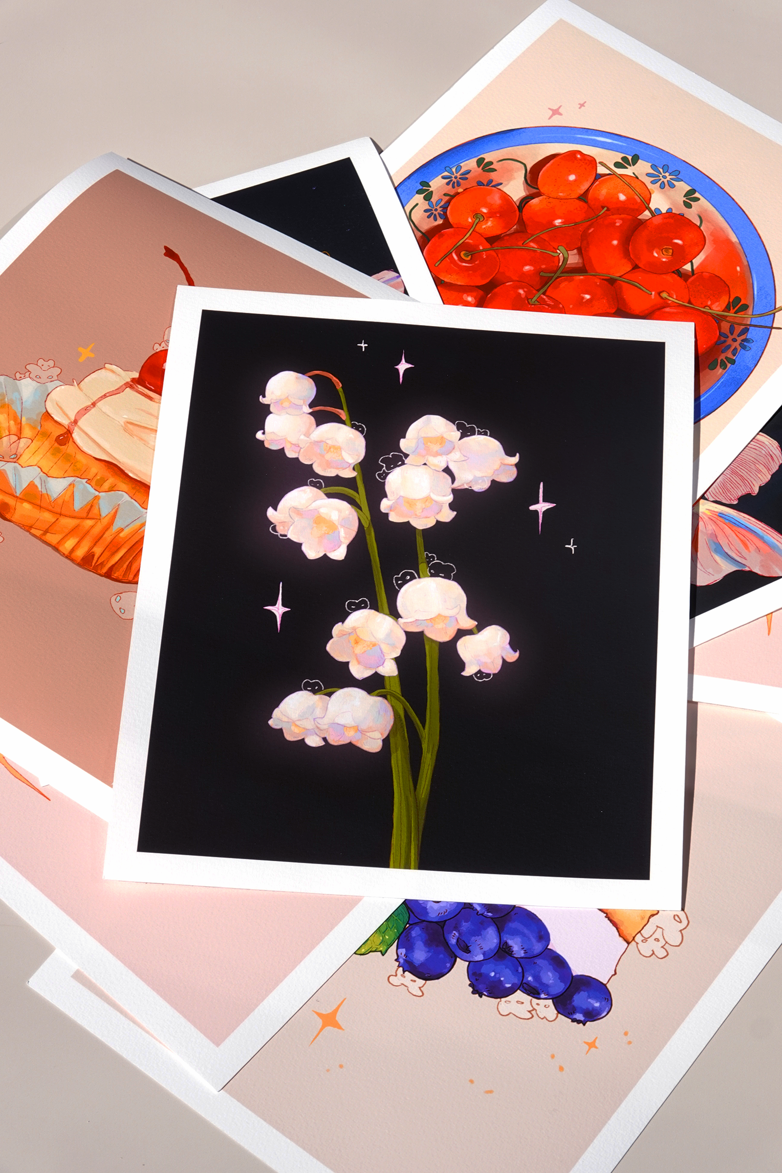 Stacked fine art prints featuring illustrated desserts, fruits, and florals including lily of the valley, cherries, blueberry cheesecake, goldfish, and cupcake designs — layered in a dynamic flat lay with visible textured paper and soft natural lighting, ideal for showcasing cohesive art print collections