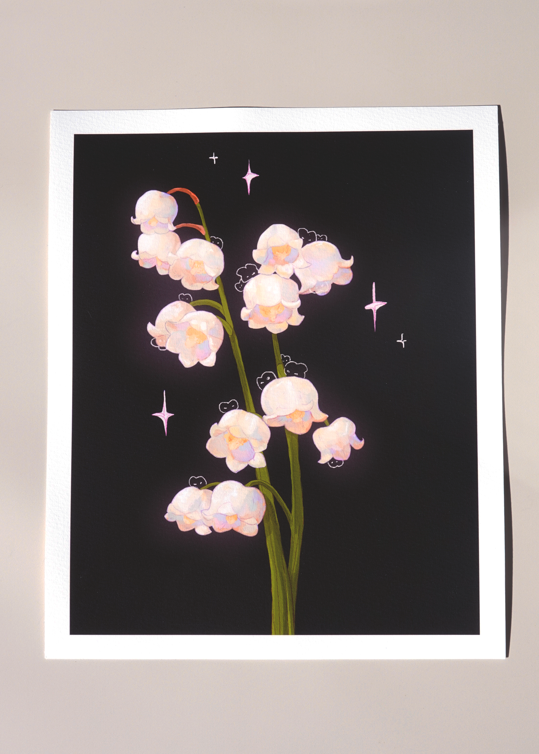 Lily of the valley fine art print on textured white paper featuring soft white bell-shaped flowers with hidden bunnies and pastel sparkles, set against a deep black background — minimal flat lay under natural light for product display or gallery wall inspiration