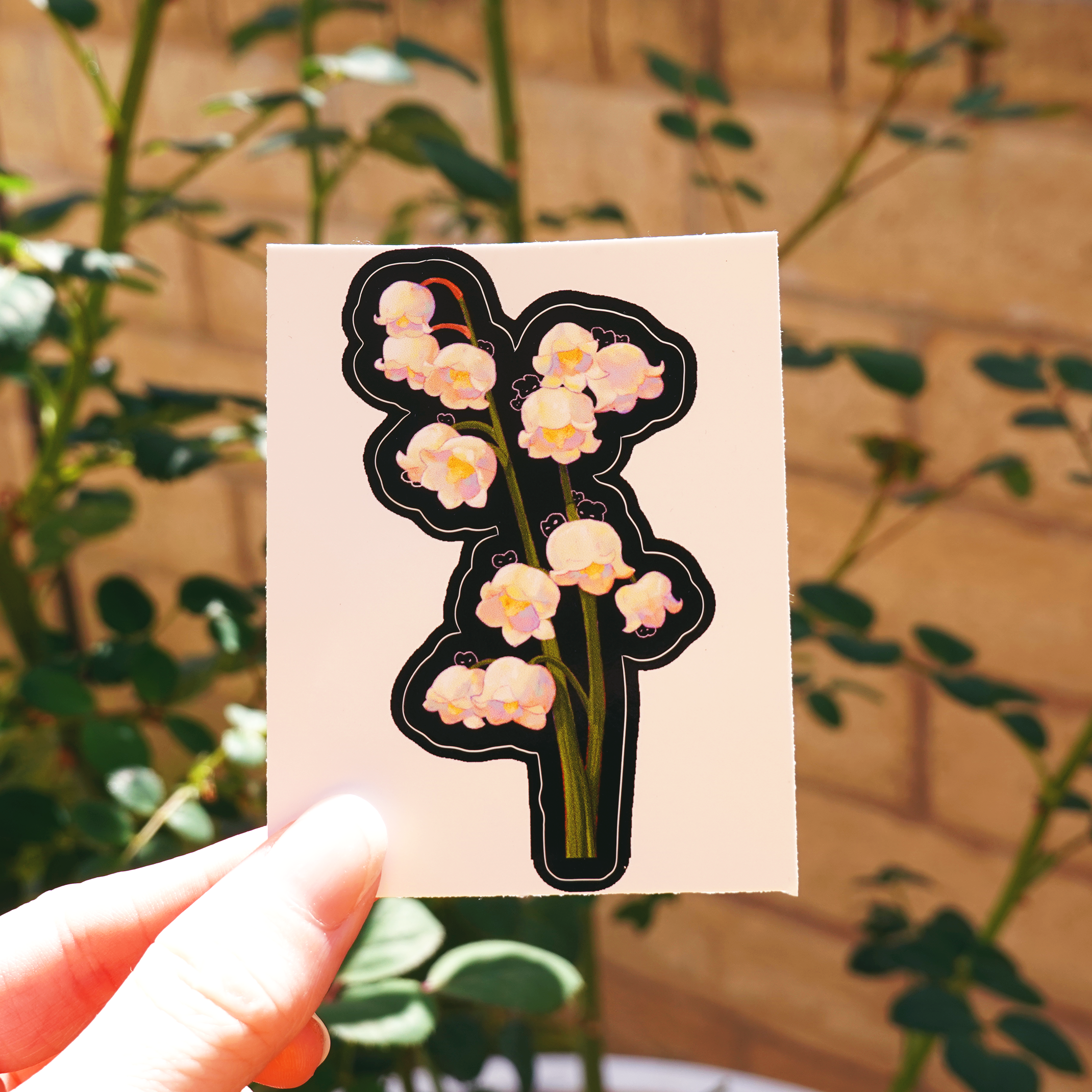 Lily of the valley vinyl sticker with hidden cute bunnies, held in hand outdoors in front of garden greenery and brick wall — glossy black die-cut floral sticker with pastel white bell-shaped flowers, perfect for water bottles, laptops, or nature-themed journaling