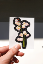 Lily of the valley floral vinyl sticker on backing card held in hand under sunlight, featuring soft pastel bell-shaped flowers with hidden cute bunny characters on a black die-cut outline — aesthetic botanical stationery for journaling, laptops, or water bottles