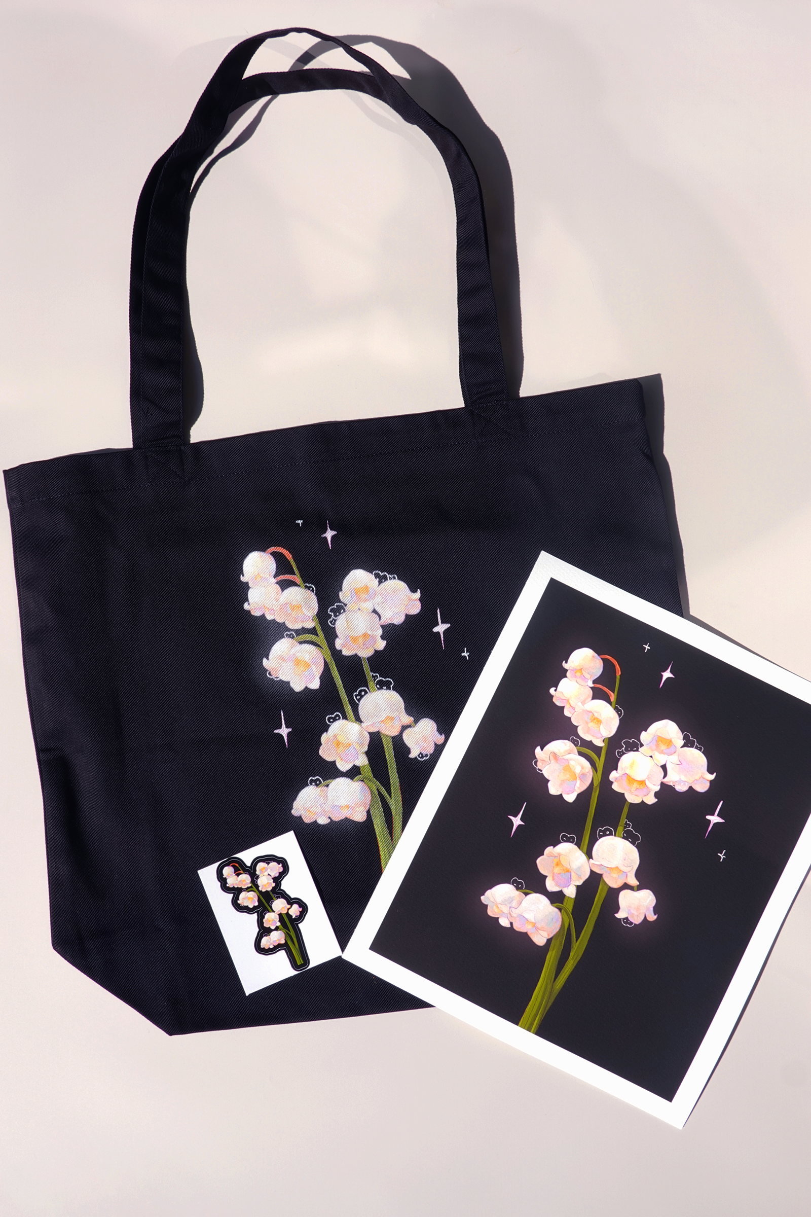 Matching lily of the valley themed art set including a black tote bag, glossy vinyl sticker, and fine art print — all featuring soft white bell-shaped flowers with hidden bunnies and pastel sparkles, arranged on a light background under natural light for an aesthetic flat lay