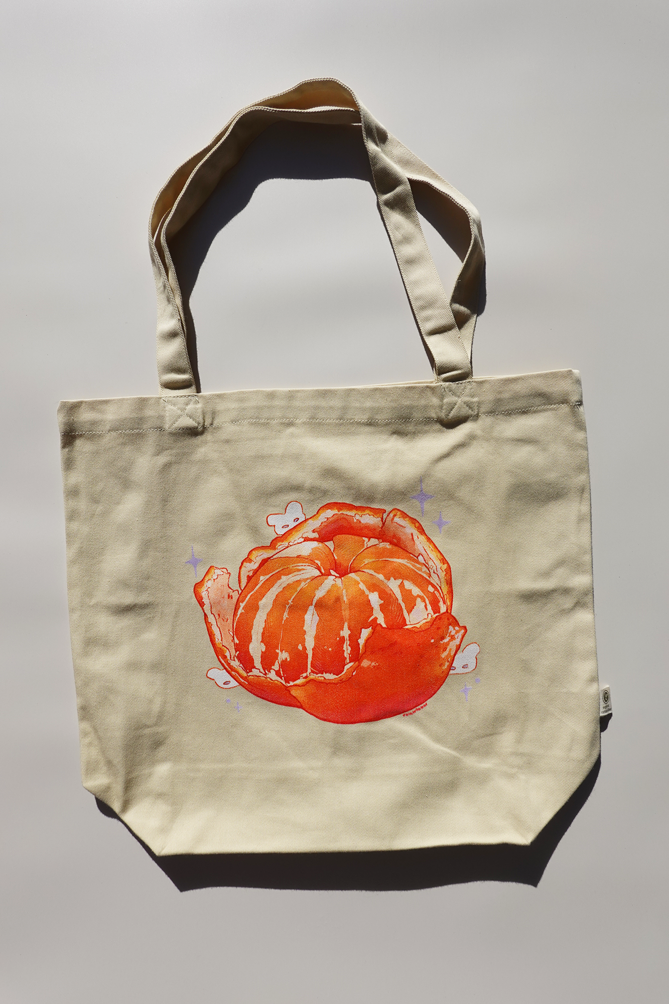 Beige canvas tote bag featuring an illustration of a peeled mandarin orange with whimsical white bunnies and purple sparkles, photographed under bright natural light.