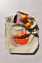 Beige canvas tote bag featuring an illustration of a peeled mandarin orange with whimsical white bunny figurines, styled with oranges and a floral book inside the bag, photographed in natural light.