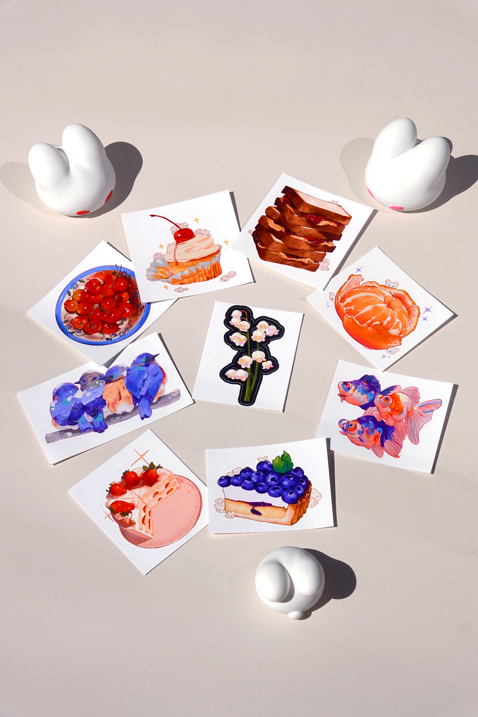 Flat lay of colorful food and animal-themed art stickers on a beige background, including blueberry cheesecake, cherry shortcake, orange slices, goldfish, bluebirds, and lily of the valley flower designs — surrounded by white bunny-shaped figures for aesthetic stationery display